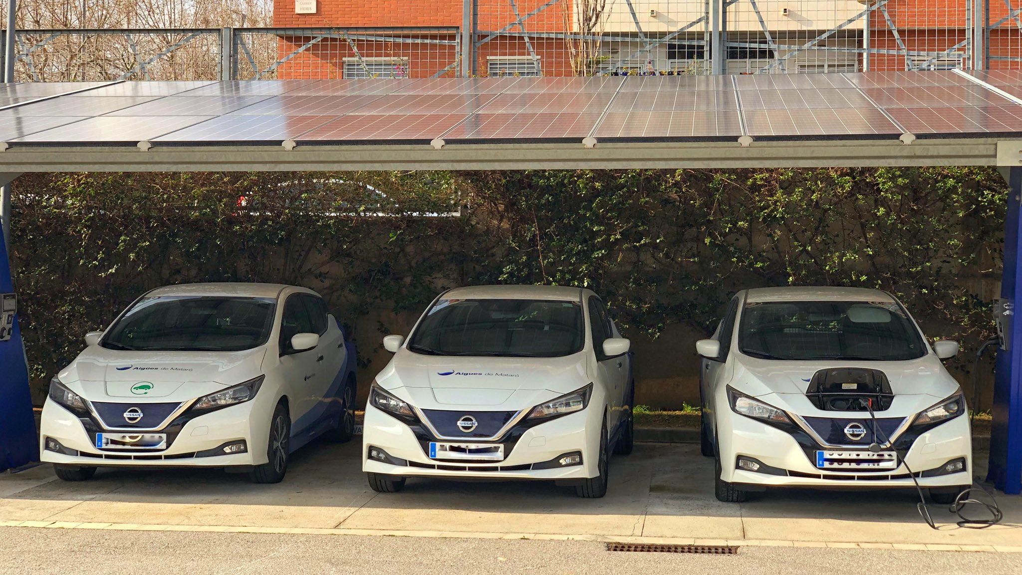 Tres vehicles elèctris de color blanc, d'Aigües de Mataró sota una coberta amb plaques fotovoltaiques per carregar-los