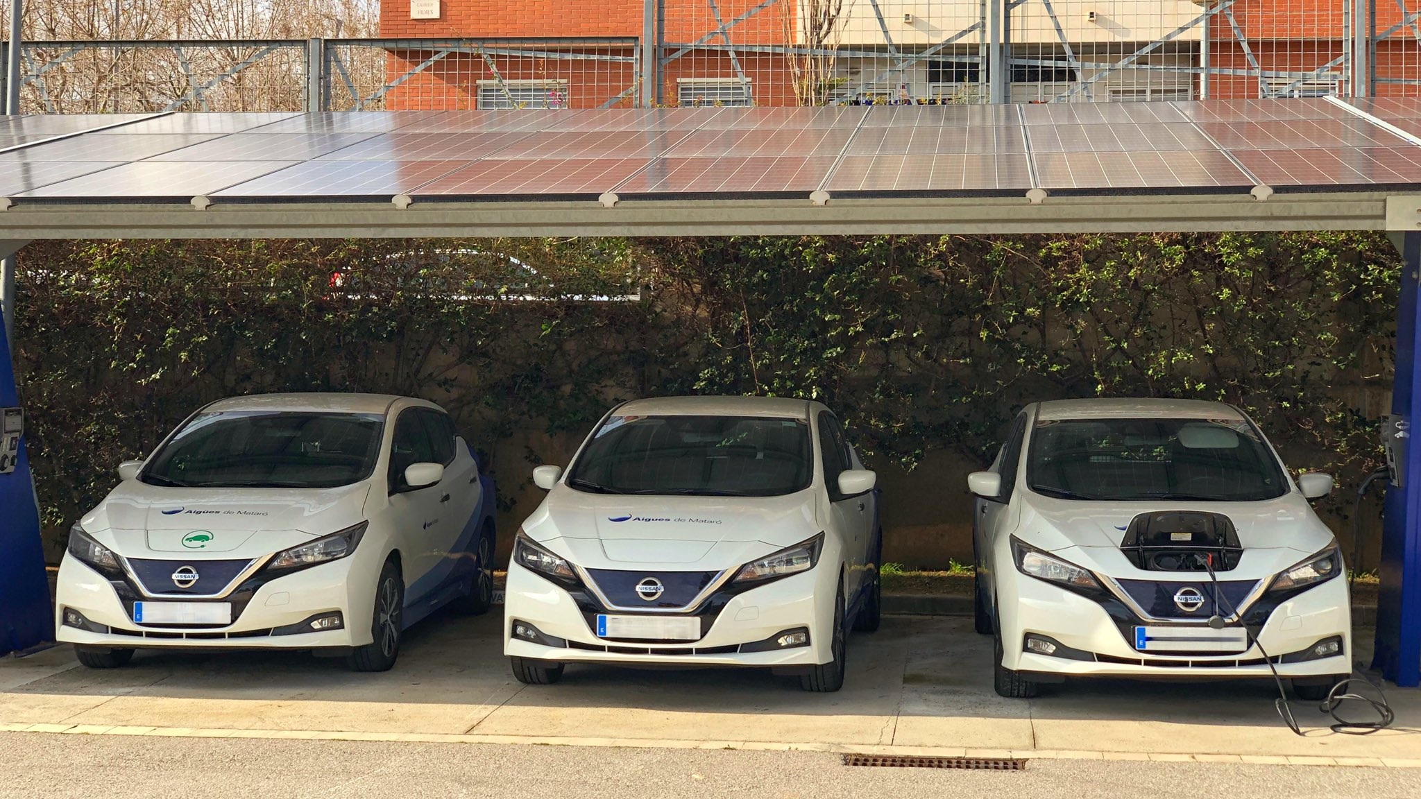 Tres vehicles elèctris de color blanc, d'Aigües de Mataró sota una coberta amb plaques fotovoltaiques per carregar-los